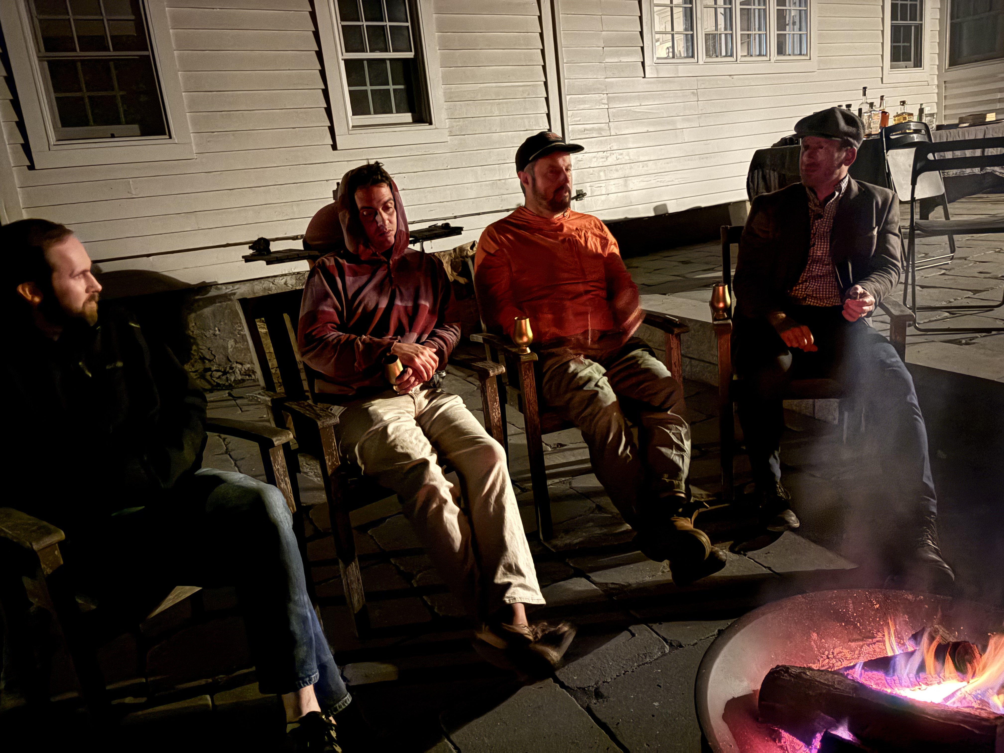 Four men gathered around an outdoor fire pit at night, enjoying cigars and whiskey.